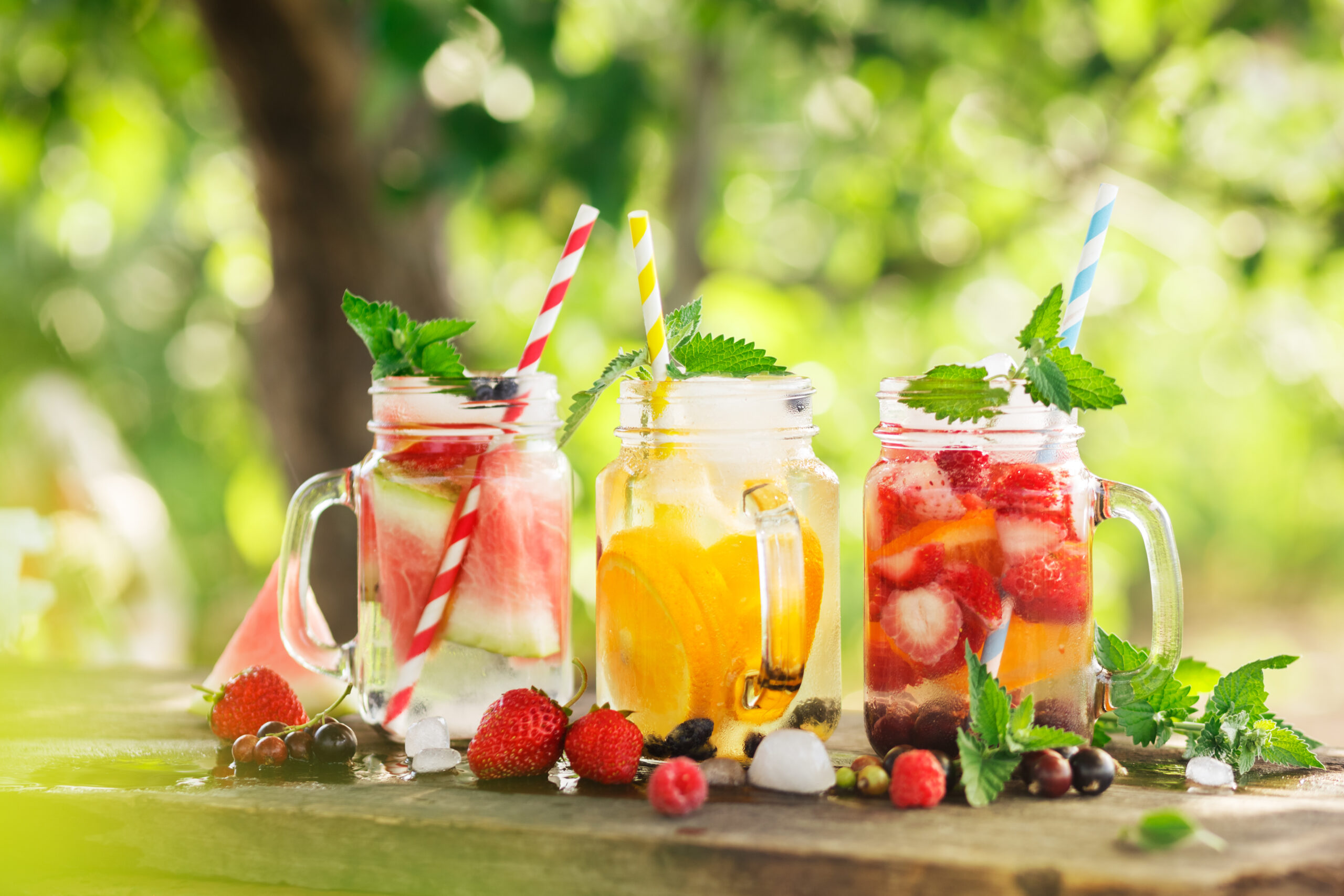 Three mason jar beverages filled with colorful fruit-infused water, garnished with berries and mint leaves. Each jar has a striped straw: red, yellow, and blue, respectively. The jars sit on a wooden surface with a blurred green, outdoorsy background.