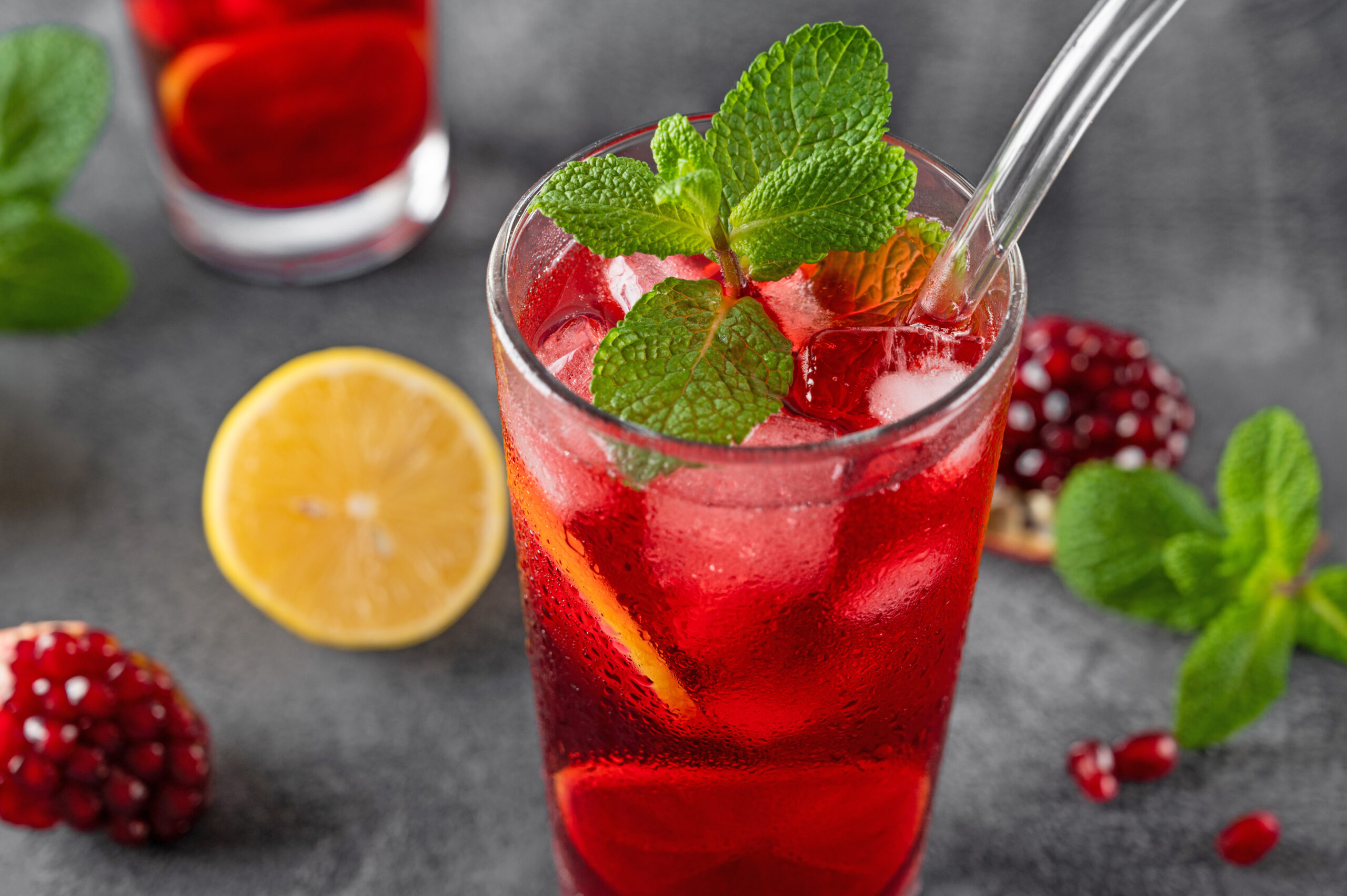 A refreshing red beverage served in a tall glass with ice, lemon slices, and garnished with fresh mint leaves. A straw is placed in the glass. In the background, there are lemons, mint leaves, and pomegranate seeds on a gray surface.