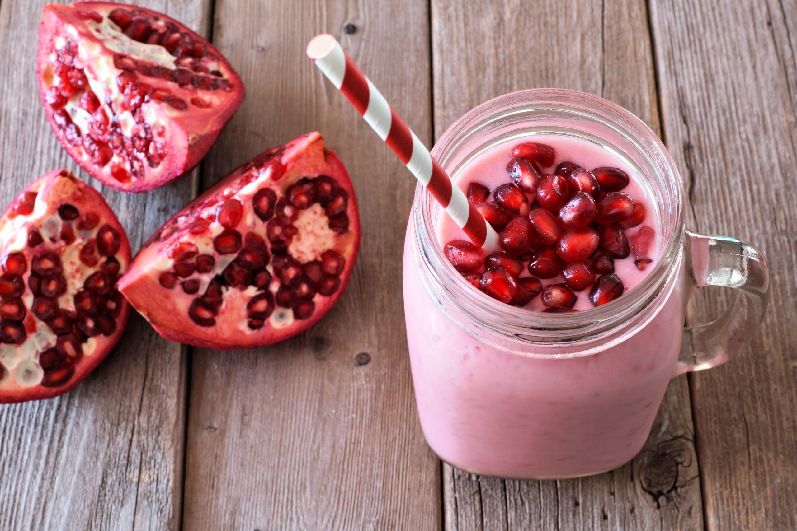 A mason jar filled with pink pomegranate frappe, topped with pomegranate seeds, and a red-and-white striped straw. Beside the jar, there are three halves of a pomegranate on a wooden table.